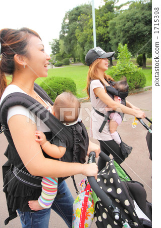 走在公園的兩個年輕媽媽通過推擠嬰兒推車 走在公園的兩個年輕媽媽通過推擠嬰兒推車 1715398