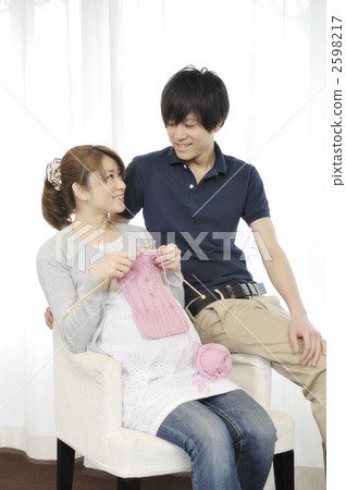 A pregnant woman knitting at the window side and a husband looking at it 2598217