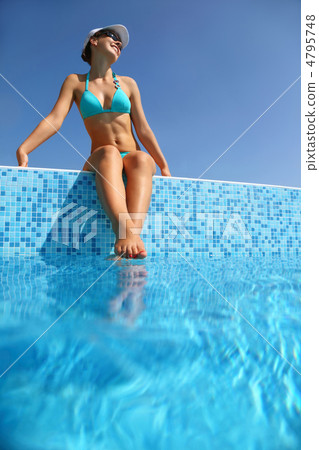 Woman in  blue swimming suit sits on verge of pool and becomes t 4795748