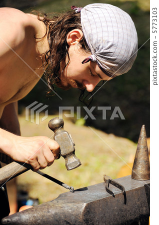 Young blacksmith hammering hot iron on anvil Young blacksmith hammering hot iron on anvil 5773103