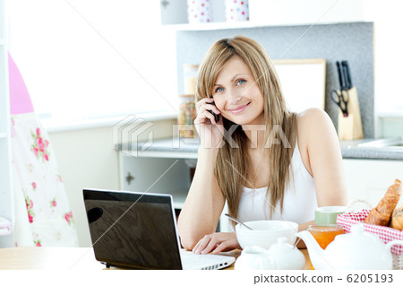 Caucasian woman using a laptop and a phone in the kitchen 6205193