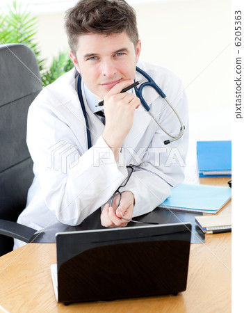 Smiling male doctor using a laptop sitting at his desk Smiling male doctor using a laptop sitting at his desk 6205363