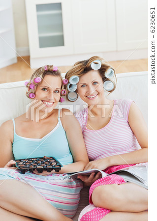 Smiling female friends with hair rollers eating chocolate readin Smiling female friends with hair rollers eating chocolate readin 6230651