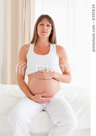 Cute pregnant female caressing her belly while sitting on a bed Cute pregnant female caressing her belly while sitting on a bed 6395634