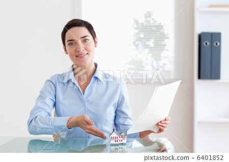 Short-haired Woman holding papers pointing at a model house 6401852