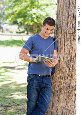 Male student leaning against a tree while using a touch pad 6773159
