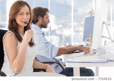 Woman biting her glasses with colleague working behind Woman biting her glasses with colleague working behind 7334018