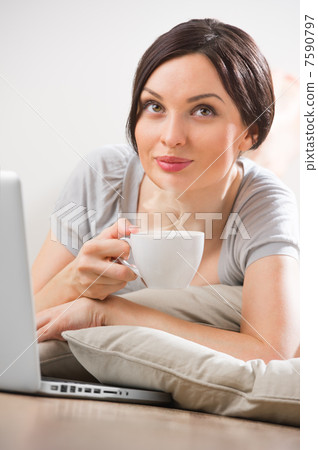 A young woman lying on the floor in front of her laptop with cup 7590797