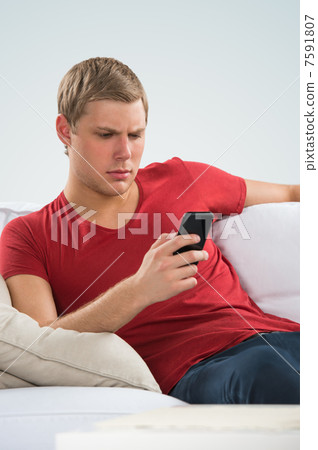 Serious young man sitting on sofa and calling with his smart pho Serious young man sitting on sofa and calling with his smart pho 7591807