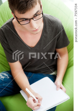 Young adult man writing in his copybook while sitting on a large Young adult man writing in his copybook while sitting on a large 7597334