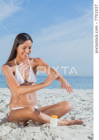 Smiling woman sitting on beach applying sunscreen 7701037