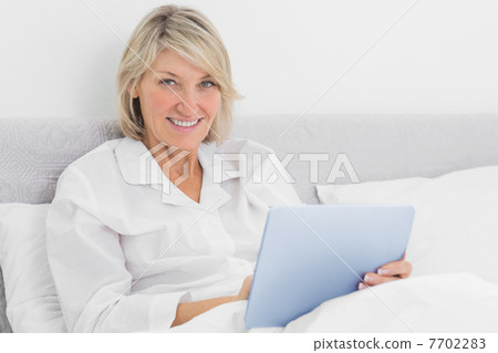 Blonde woman sitting in bed using tablet pc Blonde woman sitting in bed using tablet pc 7702283