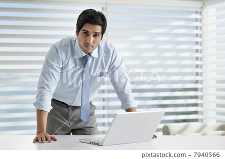 Portrait of young businessman standing by desk with laptop 7940566