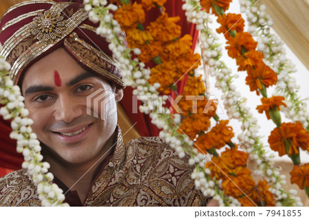 Close-up of smiling young Indian bridegroom behind wreath 7941855