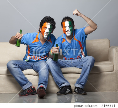 Portrait of male friends on sofa with face painted in Indian tricolor cheering while having drink at home 8166341
