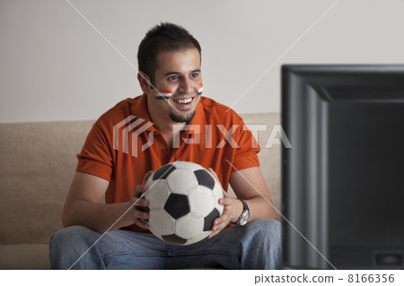 Happy young man with painted face watching television while holding soccer ball 8166356