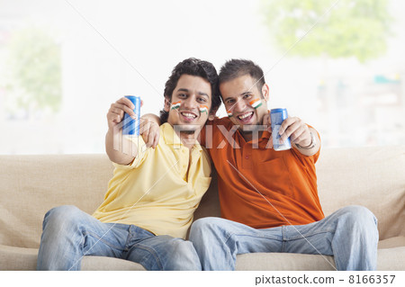 Portrait of cheerful young male friends with face painted holding out tin cans while sitting on sofa Portrait of cheerful young male friends with face painted holding out tin cans while sitting on sofa 8166357
