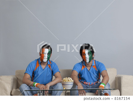 Male friends with face painted in Indian tricolor sitting on sofa with bowl of popcorn on table at home Male friends with face painted in Indian tricolor sitting on sofa with bowl of popcorn on table at home 8167154