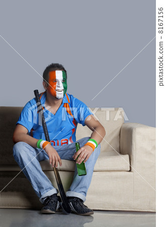 Young man with painted face in Indian tricolor sitting on sofa with hockey stick and beer bottle 8167156