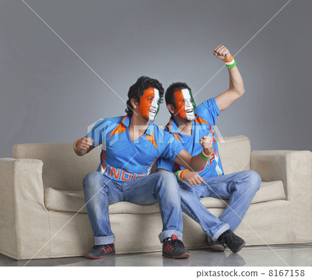 Young male friends with face painted in tricolor cheering while sitting on sofa 8167158