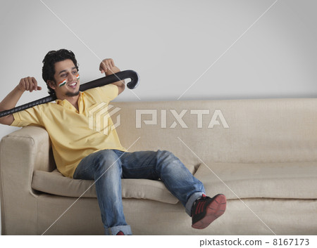 Young man in casuals on sofa looking away while holding hockey stick 8167173