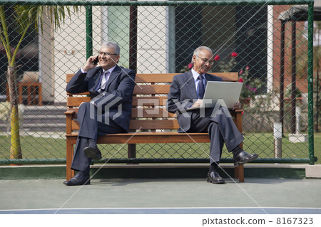 Senior businessman on phone call with other executive using laptop while sitting on bench in a tennis court 8167323