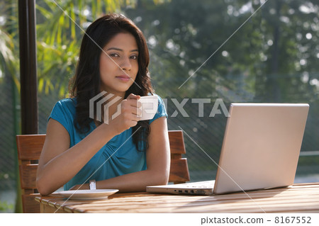 Portrait of an attractive young woman having tea while sitting by table with laptop 8167552