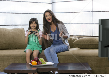 Little girl playing video game with mother sitting besides her on sofa at home 8167678