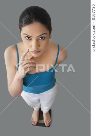 Portrait of worried young woman standing on weighing scale isolated over gray background 8167750