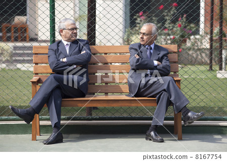 Senior male business executives staring at each other while sitting arms crossed on bench in tennis court 8167754