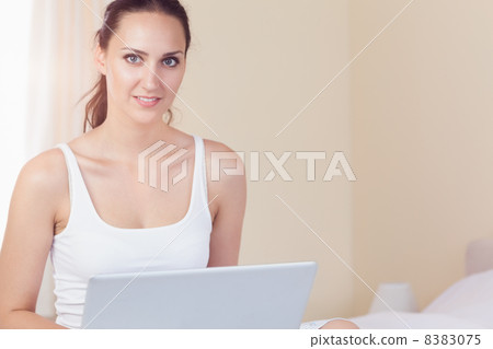 Happy brunette sitting on bed using her laptop Happy brunette sitting on bed using her laptop 8383075
