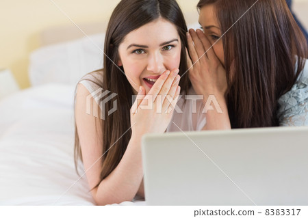 Happy girl telling a secret to her friend in front of laptop Happy girl telling a secret to her friend in front of laptop 8383317