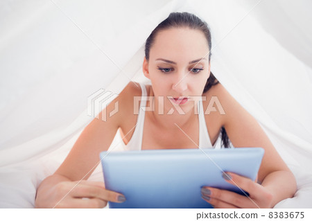 Happy brunette lying under the sheets using her tablet pc Happy brunette lying under the sheets using her tablet pc 8383675