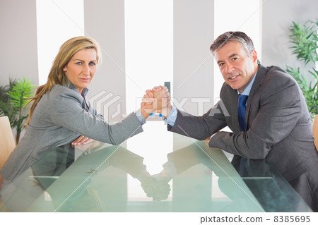 Two serious businesspeople having an arm wrestling sitting aroun Two serious businesspeople having an arm wrestling sitting aroun 8385695