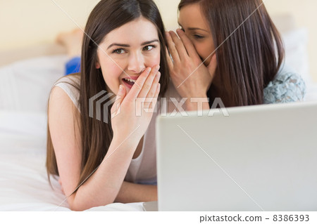 Smiling girl telling a secret to her friend in front of laptop 8386393