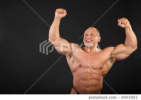 Undressed tanned happy bodybuilder raises his fists up in black 8403981