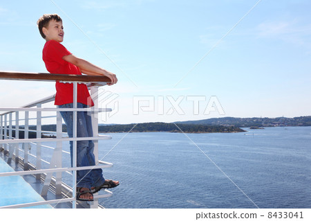 Boy stands at railing on deck of ship, looks into distance and s 8433041