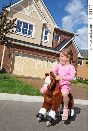 Little girl rides at toy horse near cottage at sunny summer day. 8433145