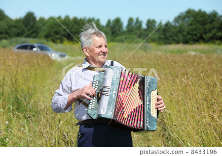 Grandfather in shirt play on accordion in field near car 8433196