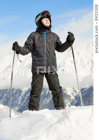 Boy in ski suit stands leaning on ski poles and looking upward, 8433308