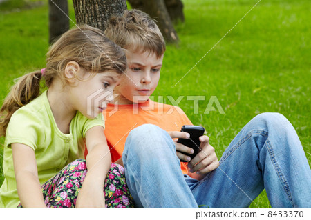 Boy in orange t-shirt sits leaning his back on trunk of tree and 8433370