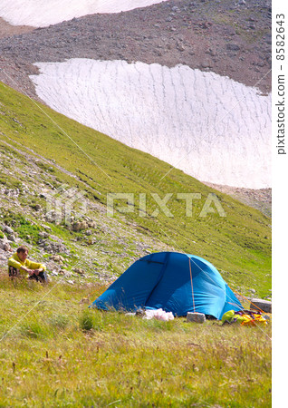 Camping Tent in Mountains with Man Hiker sitting on grass and Glacier Hillside on Background 8582643