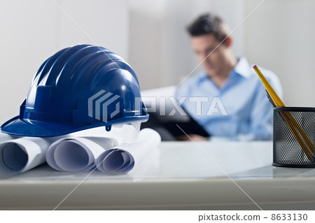 hardhat and blueprint on desk, with architect in background 8633130