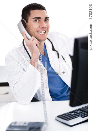 Smiling young physician sitting at his desk talking over phone 8782108