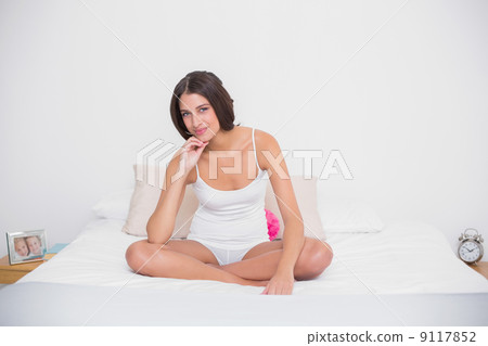 Thoughtful young brown haired model in white pajamas sitting on 9117852