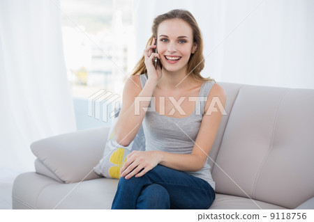 Happy young woman sitting on couch talking on smartphone 9118756