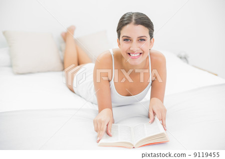 Delighted young brown haired model in white pajamas reading a bo 9119455