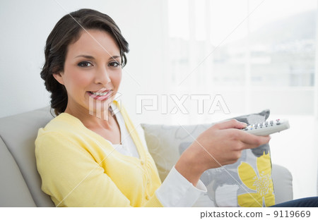 Pleased casual brunette in yellow cardigan changing tv channel Pleased casual brunette in yellow cardigan changing tv channel 9119669