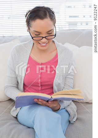 Smart woman with reading glasses sitting on sofa and reading a b 9122247