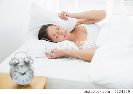 Woman covering ears with pillow and alarm clock on side table 9124539